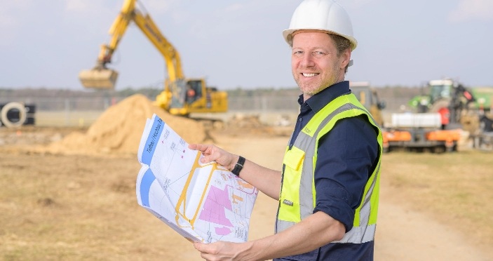 Person mit weißem Schutzhelm und gelber Warnweste hält Baupläne auf einer Baustelle mit Baggern im Hintergrund