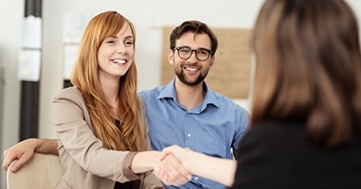 Zwei Personen sitzen an einem Tisch und geben einer dritten Person die Hand