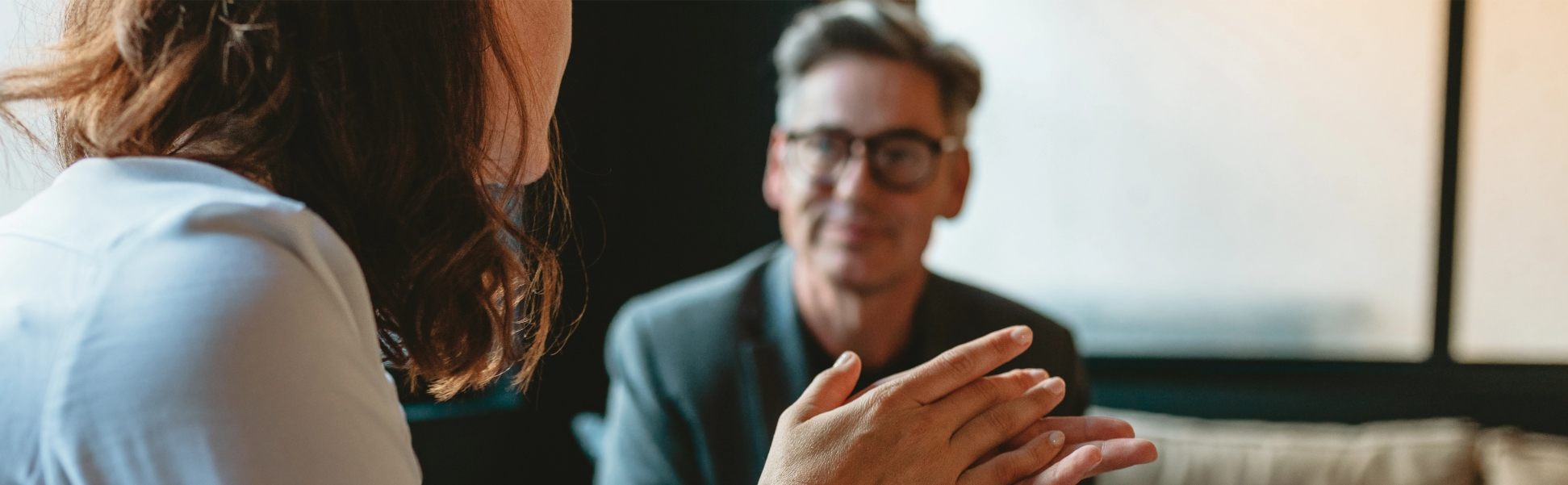 Zwei Personen im Gespräch, eine gestikuliert mit den Händen, beide sitzen an einem Tisch vor einem Fenster